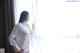 A woman in a white shirt and panties standing by a window.