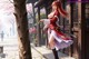 A woman in traditional attire stands near a tree, holding a sword, with cherry blossoms and a wooden building in the background.