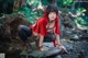 A woman in a red hoodie crouching on a rock in the woods.